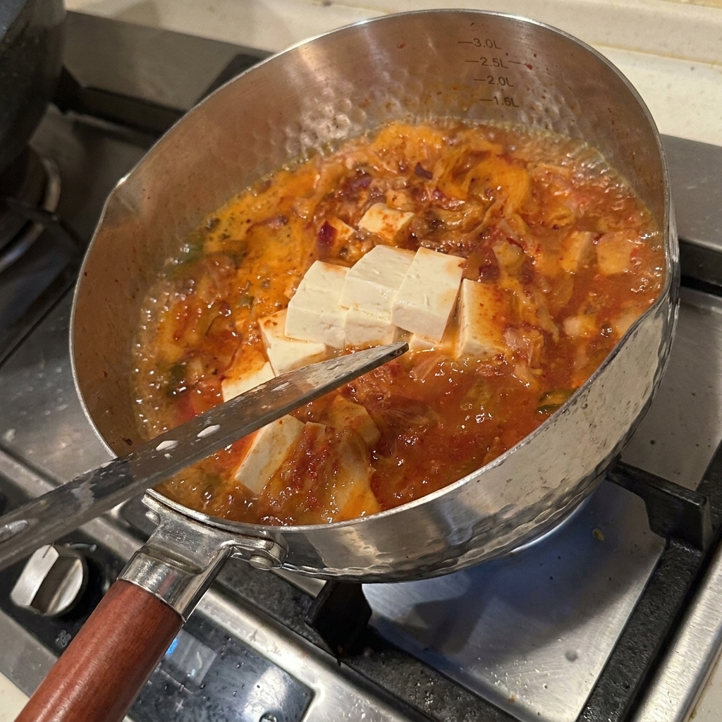Verschiedene frische Zutaten wie Tofuwürfel, Enoki-Pilze und Kimchi köcheln in einer würzigen, rot gefärbten Brühe.