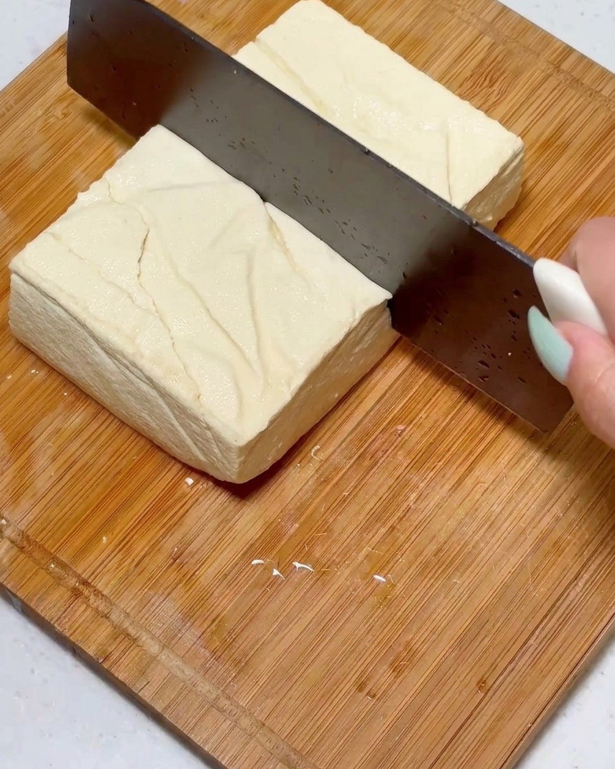 Ein fester Tofu-Block wird mit einem großen Messer auf einer Holzoberfläche in gleichmäßige Stücke geschnitten.