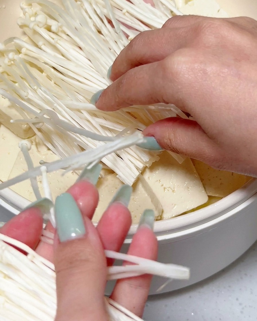 Hände platzieren ein großes Büschel frischer weißer Enoki-Pilze auf den geschichteten Zutaten in einem Kochtopf.