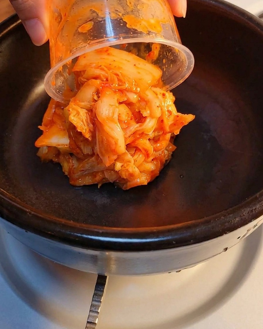 Fermentiertes Kimchi und zerkleinertes Frühstücksfleisch werden in einem Steintopf geschichtet.
