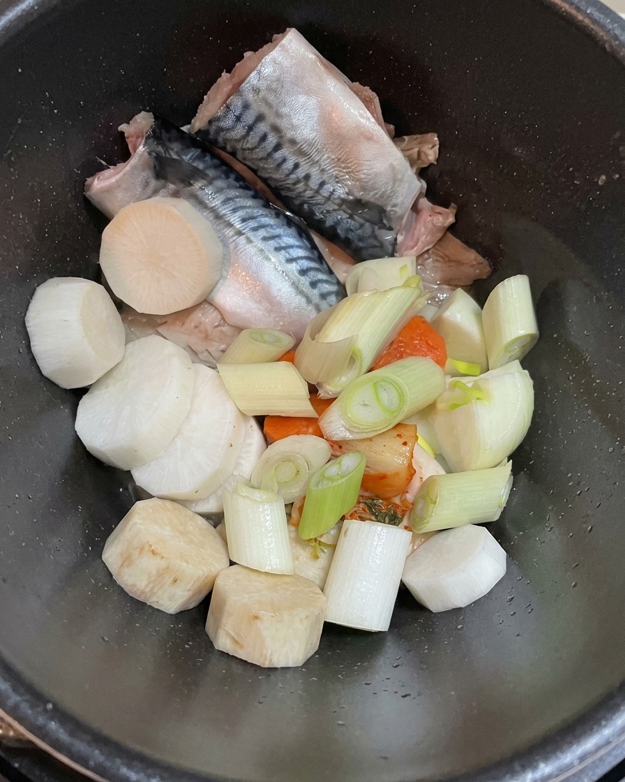 Blick von oben in einen dunklen Topf mit rohen Makrelenfilets, stückigem weißem Rettich und frisch geschnittenen Frühlingszwiebeln.