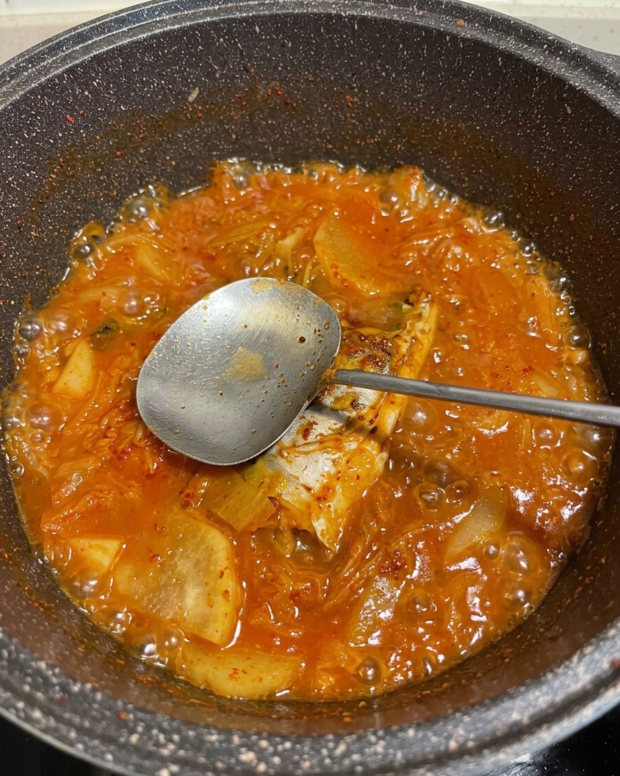 Ein kräftig roter, sprudelnder Fischeintopf mit Gemüse im Topf, ein Metalllöffel hebt ein Fischstück an.