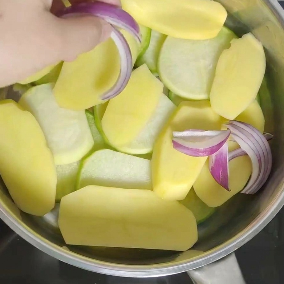 Eine Hand gibt frische rote Zwiebelstücke in den Topf auf die Kartoffelschicht.