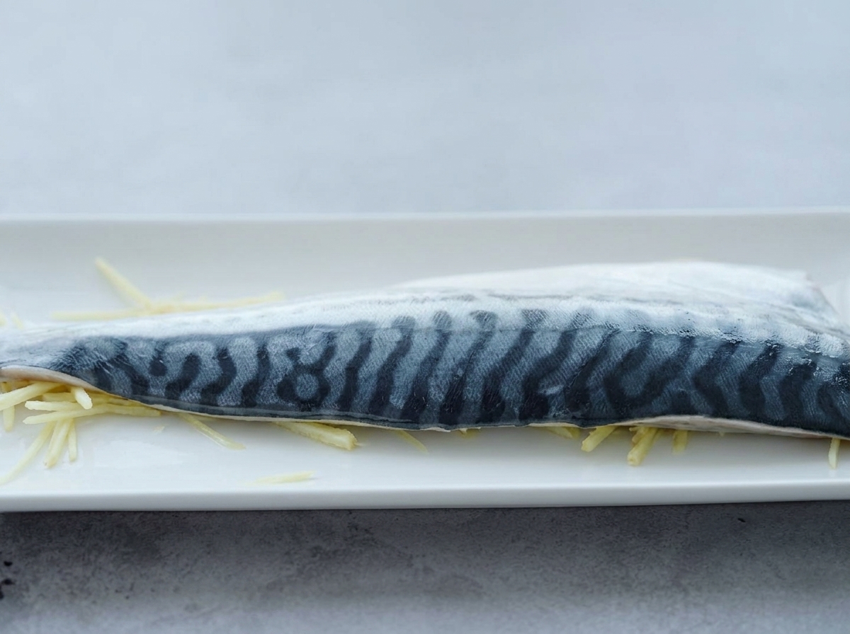 Ein rohes Makrelenfilet liegt auf dünnen Ingwerstreifen, während Koch-Sake darüber gegossen wird.
