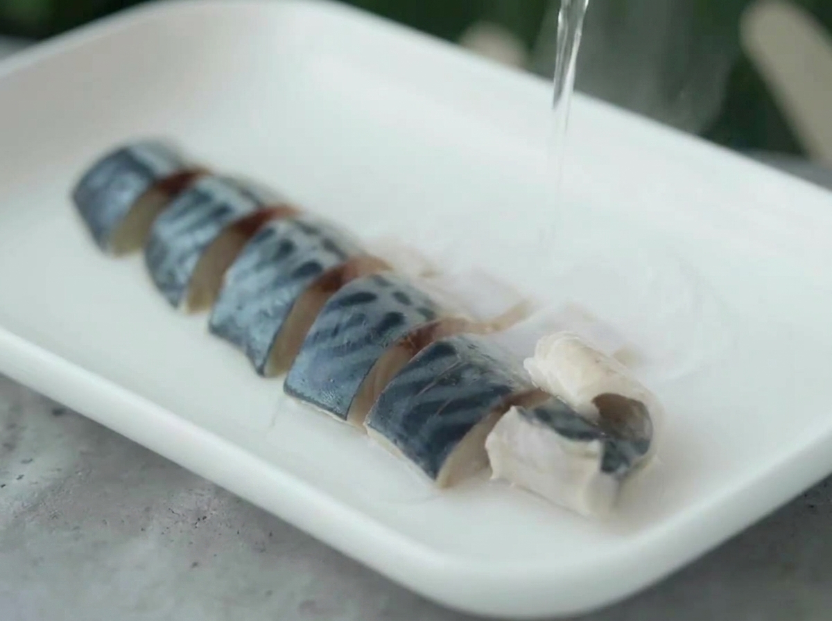 Heißes Wasser wird aus einem Wasserkocher über die angeordneten Makrelenstücke auf einem weißen Teller gegossen.