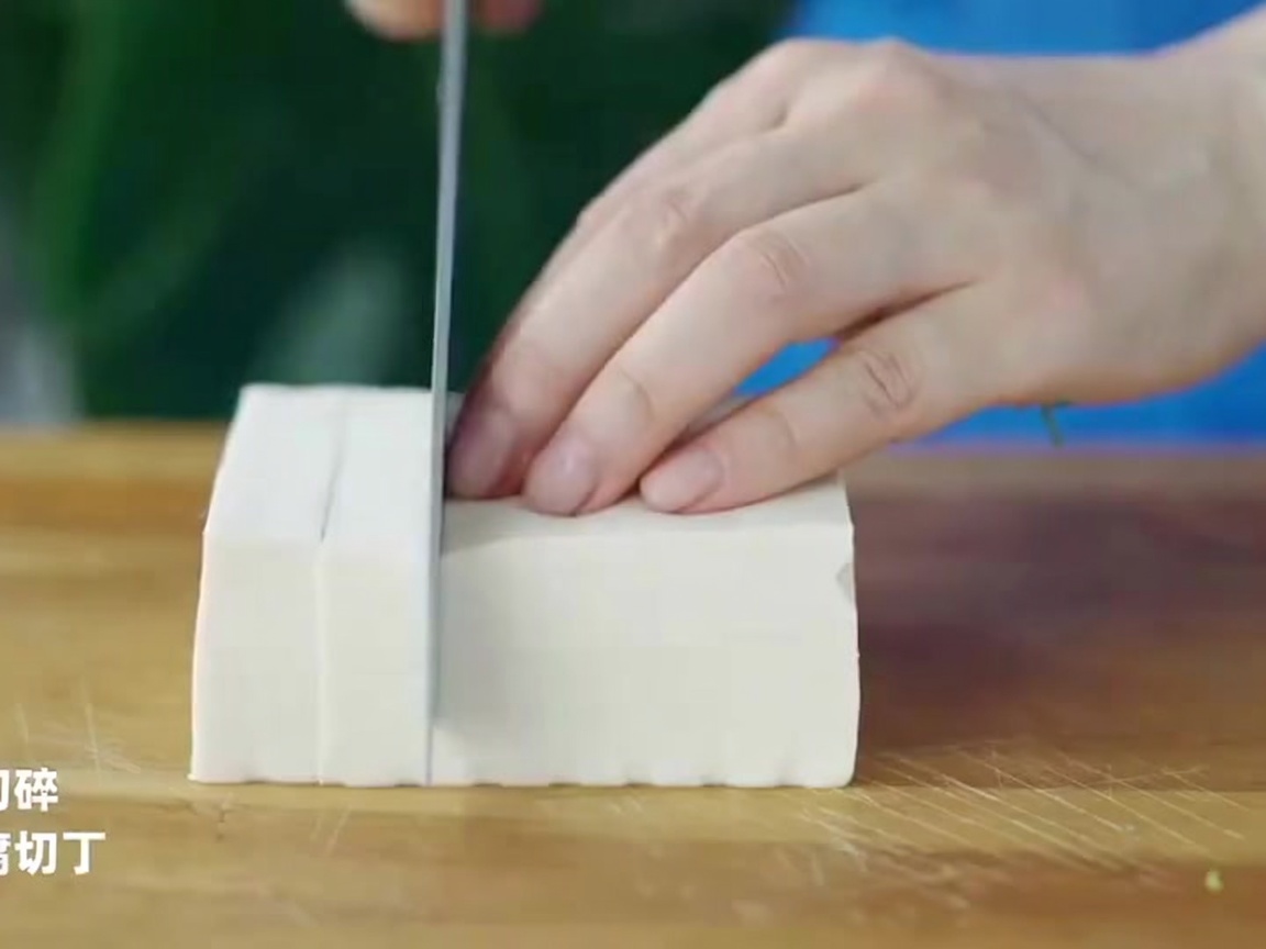 Ein Kochmesser schneidet einen Block weißen Tofu auf einer Holzoberfläche in präzise, gleichmäßige Würfel.