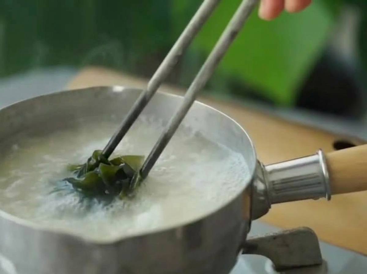 Rehydrierte grüne Wakame wird mit Essstäbchen in die köchelnde Miso-Suppe gegeben.