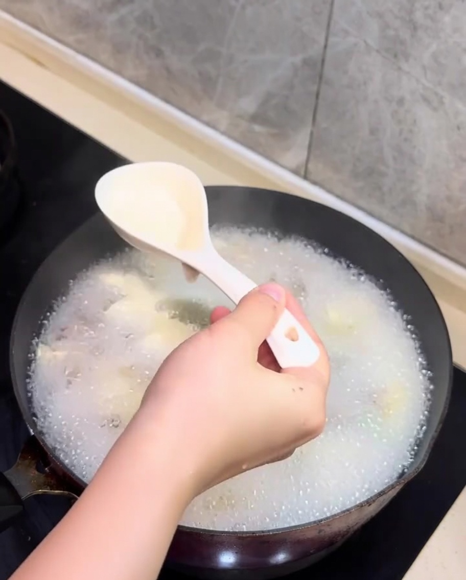 Ein Löffel schöpft weißen Schaum von der Oberfläche der köchelnden Muschel-Tofu-Suppe.