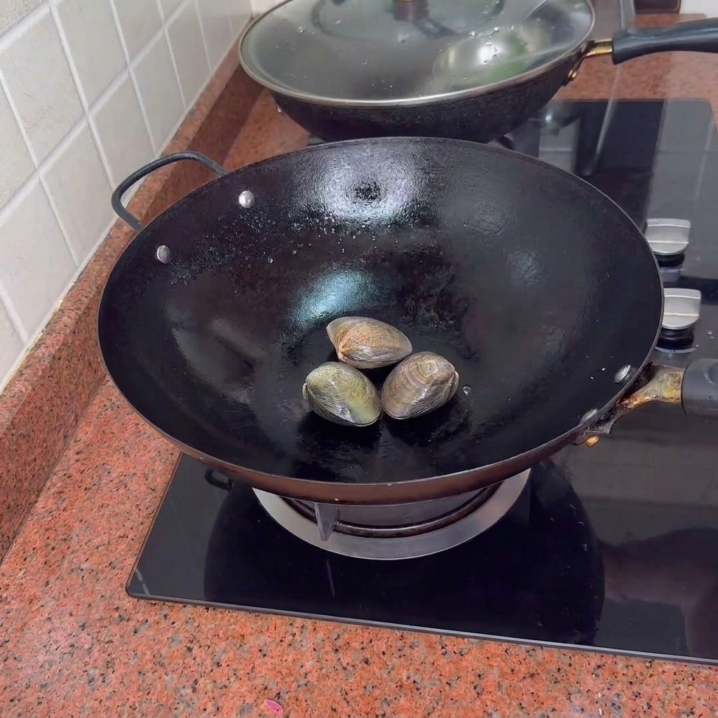 Drei geschlossene Venusmuscheln in einem schwarzen Wok auf einem Herd.