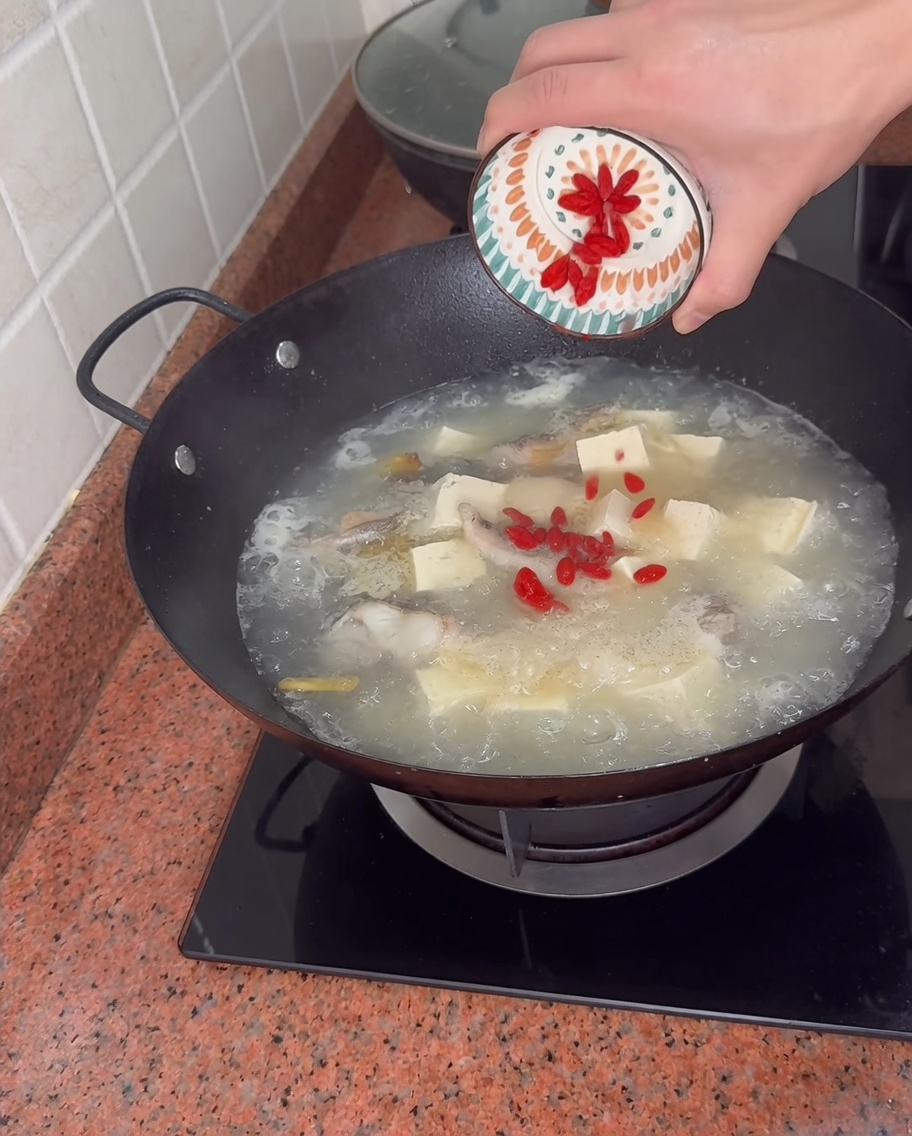 Rote Goji-Beeren aus einer kleinen gemusterten Schale in einen sprudelnden Wok mit Fisch- und Tofusuppe geben.