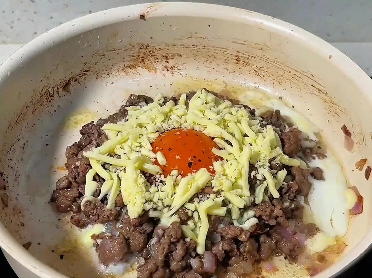 Geriebener Käse bedeckt das Rinderhack und das Ei, die Pfanne ist mit einem Deckel abgedeckt.