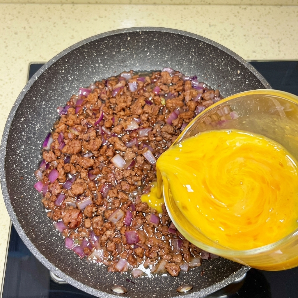 Verquirlte Eier werden aus einem Messbecher über das Fleisch und die Zwiebeln in die Pfanne gegossen.