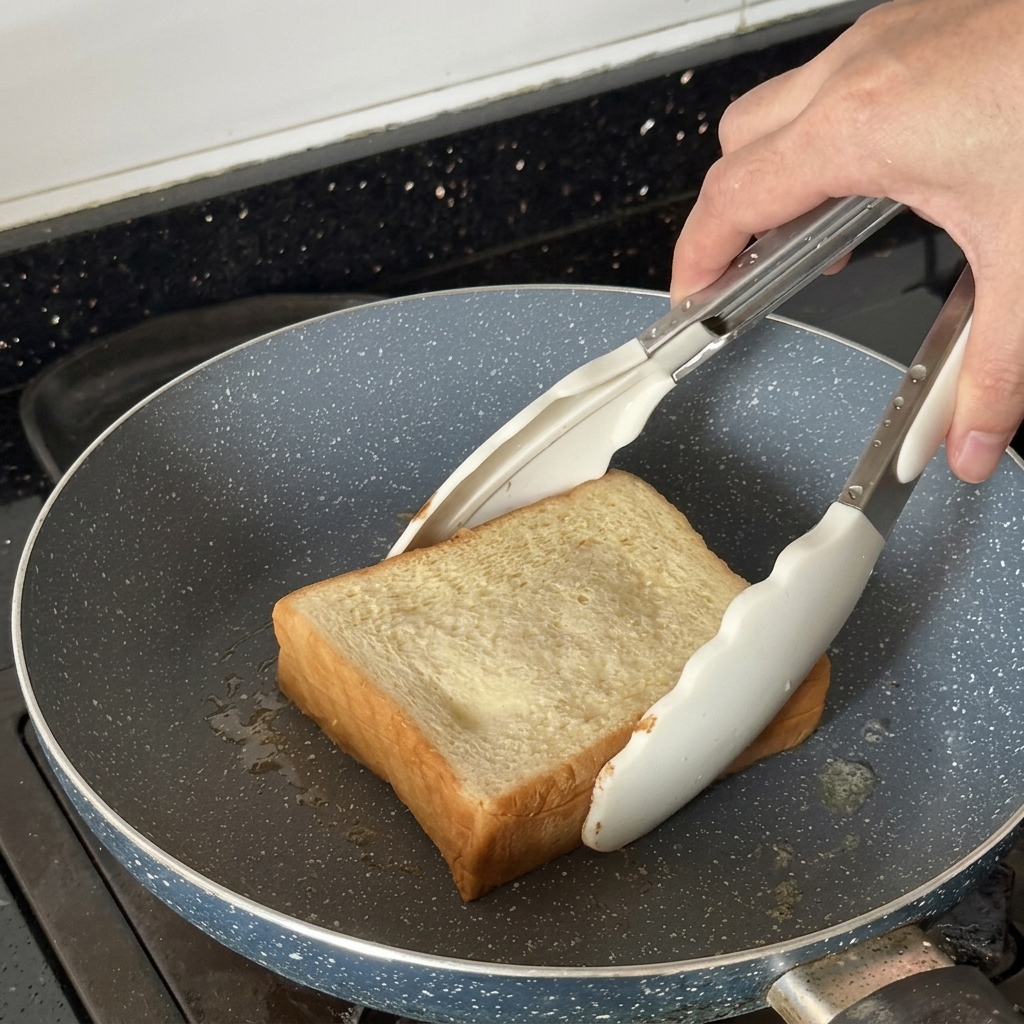 Eine dicke Scheibe Brot wird mit einer weißen Zange in einer Pfanne geröstet.