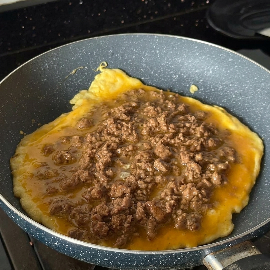 Ein rundes Omelett in einer Pfanne, großzügig belegt mit gebratenem Hackfleisch.