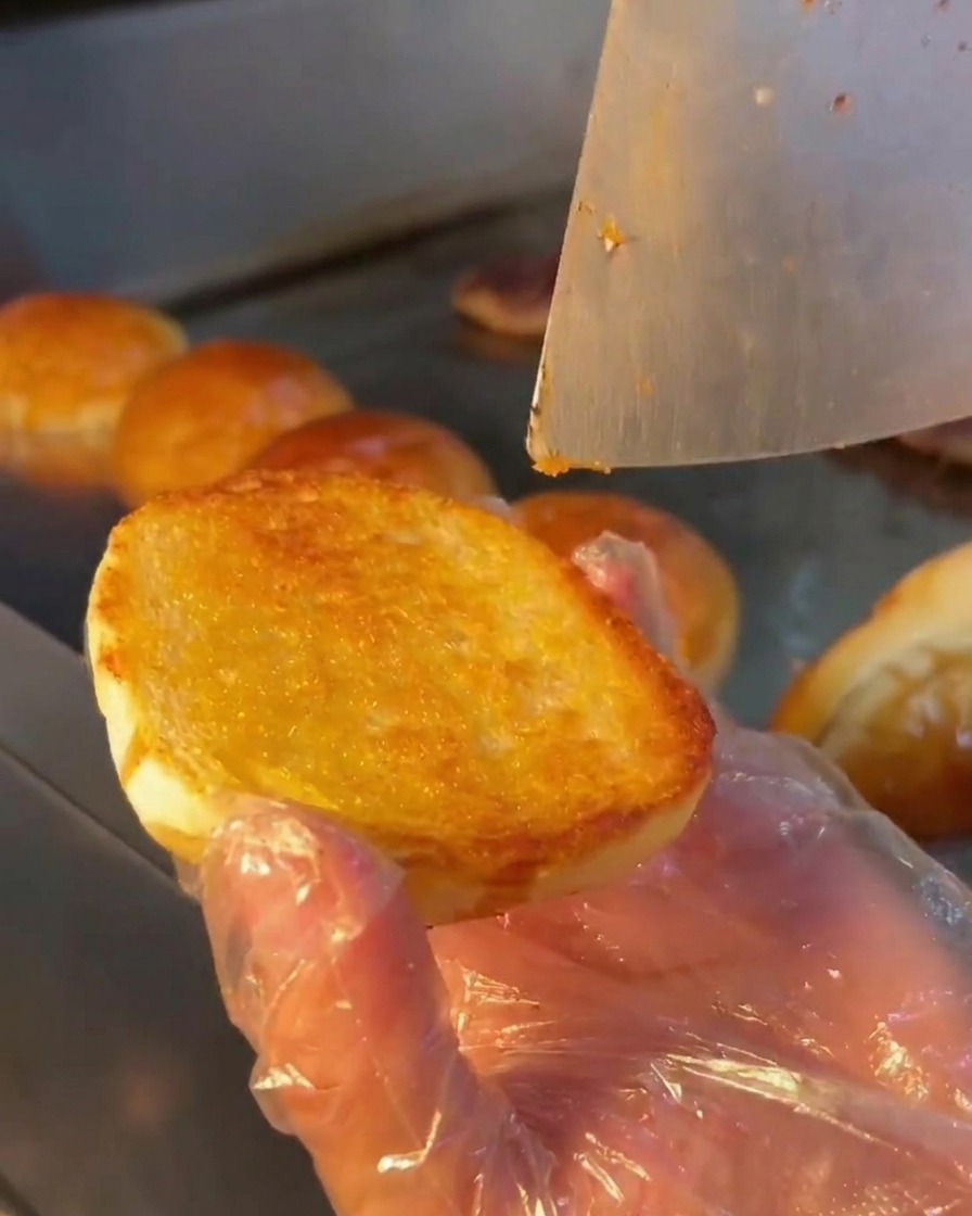 Ein Metalllöffel verteilt eine cremige weiße Sauce auf der getoasteten Oberfläche eines Burgerbrötchens.