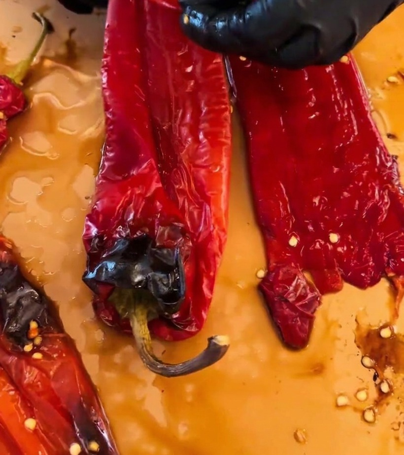 Hände mit schwarzen Handschuhen ziehen die verkohlte Haut von gerösteten roten Chilis auf einem Schneidebrett ab.