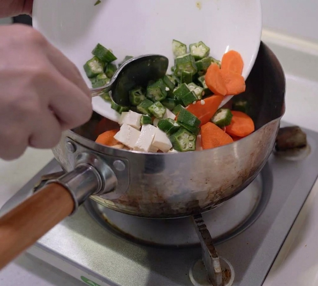 Vorbereitete Okra, Karotten, Pilze und Tofu, die in einen Edelstahltopf gegeben werden.