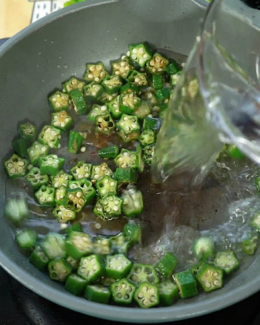 Wasser wird in eine Pfanne mit angebratenen Okraschoten gegossen.