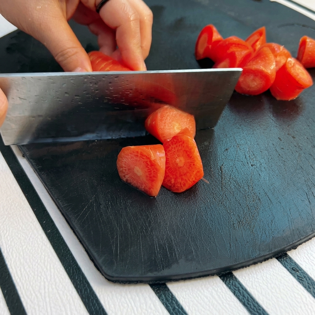 Eine Hand schneidet mit einem großen Metallbeil eine leuchtend orangefarbene Karotte in grobe Roll-Cut-Stücke auf einem schwarzen Brett.