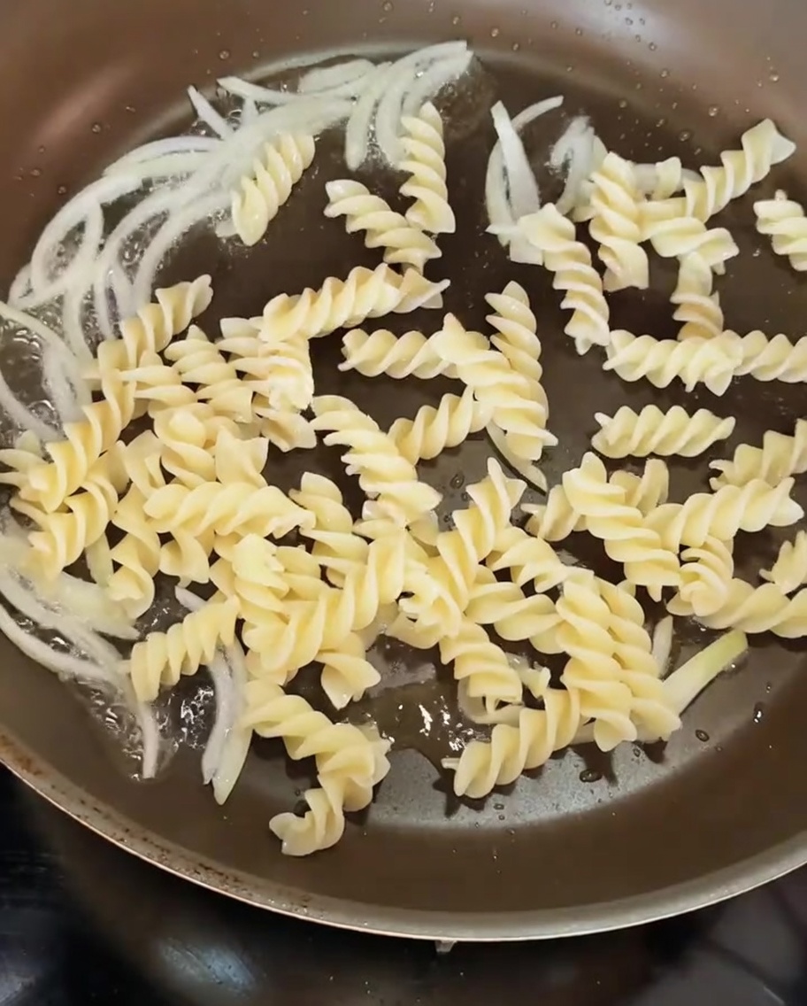 Gekochte Spiralnudeln, die in eine Pfanne mit Zwiebelringen gegeben werden.