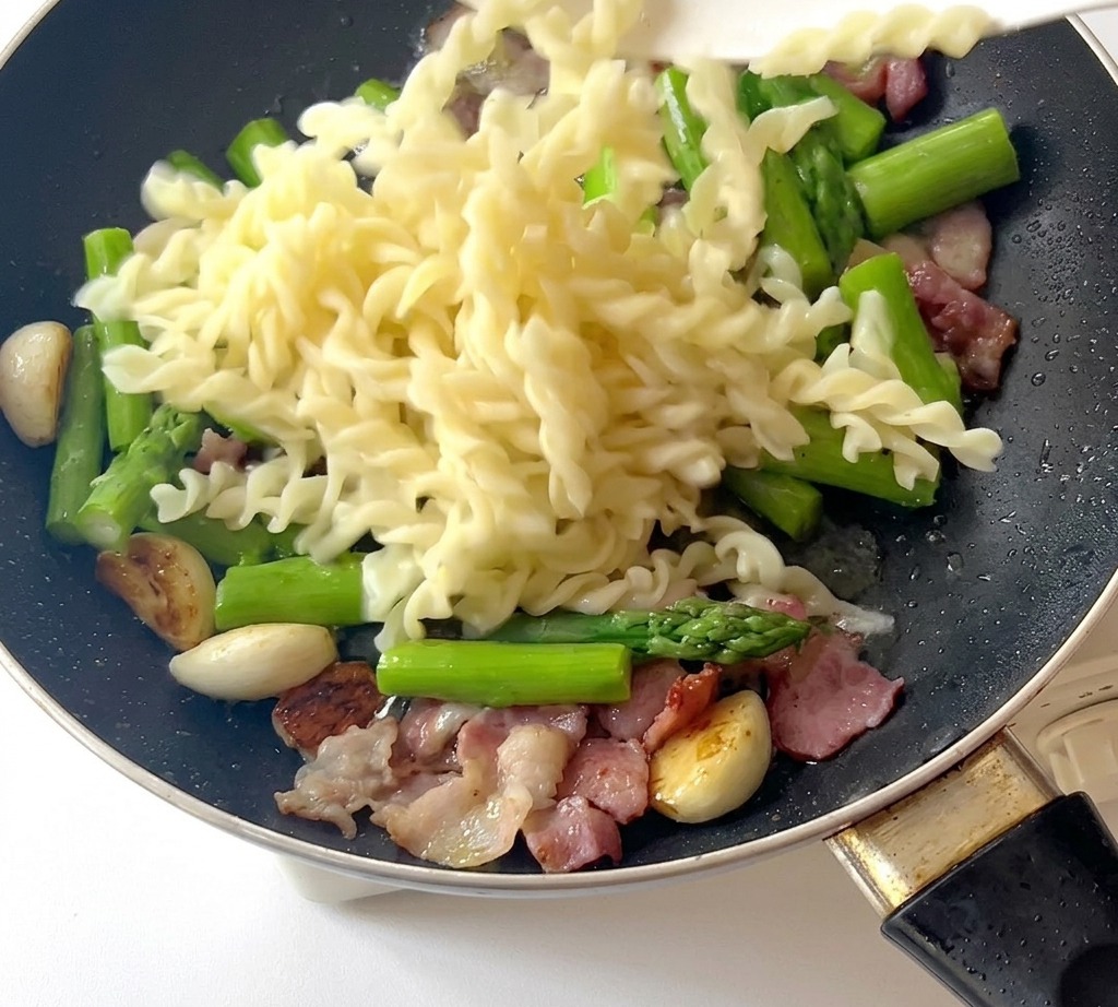 Gekochte Fusilli Nudeln werden in die Pfanne zu Spargel und Speck gegeben.