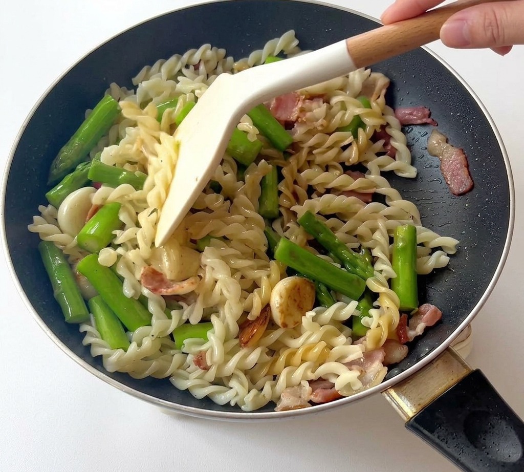 Ein Pfannenwender vermengt Fusilli, Spargel und Speck in der Pfanne.