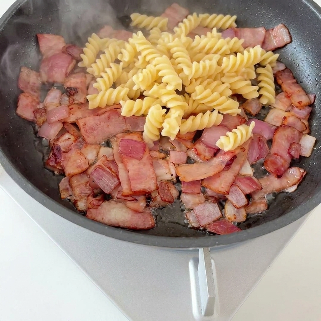 Fusilli-Pasta und leuchtend grüne Spargelstücke liegen auf einer Basis aus knusprigem Speck und karamellisierten Zwiebeln in einer Pfanne.