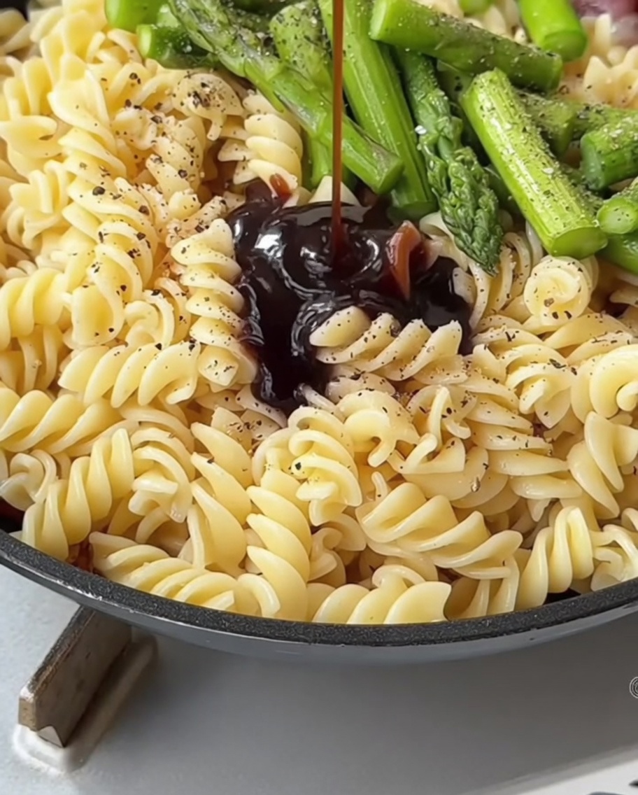 Eine dunkle Saucenmischung wird aus einem Löffel über die Pasta und den Spargel in die Pfanne gegossen.
