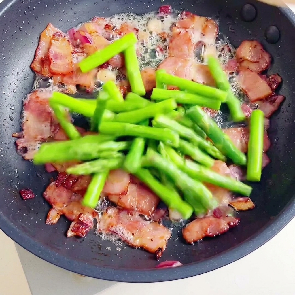 Knuspriger Speck, grüner Spargel und gekochte Fusilli in einer Pfanne.