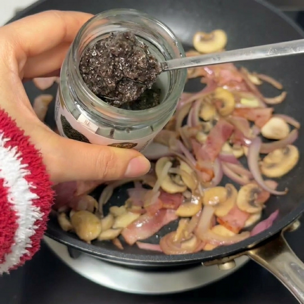 Ein Löffel schwarze Trüffelsauce wird unter die Mischung aus Speck, Zwiebeln und Pilzen gerührt.
