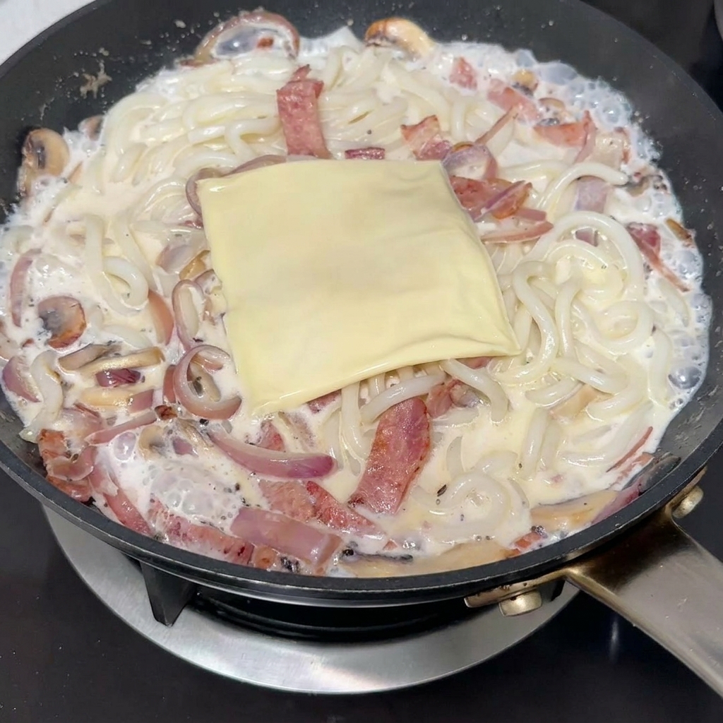 Eine Scheibe Schmelzkäse liegt auf den cremigen Udon-Nudeln in der Pfanne.
