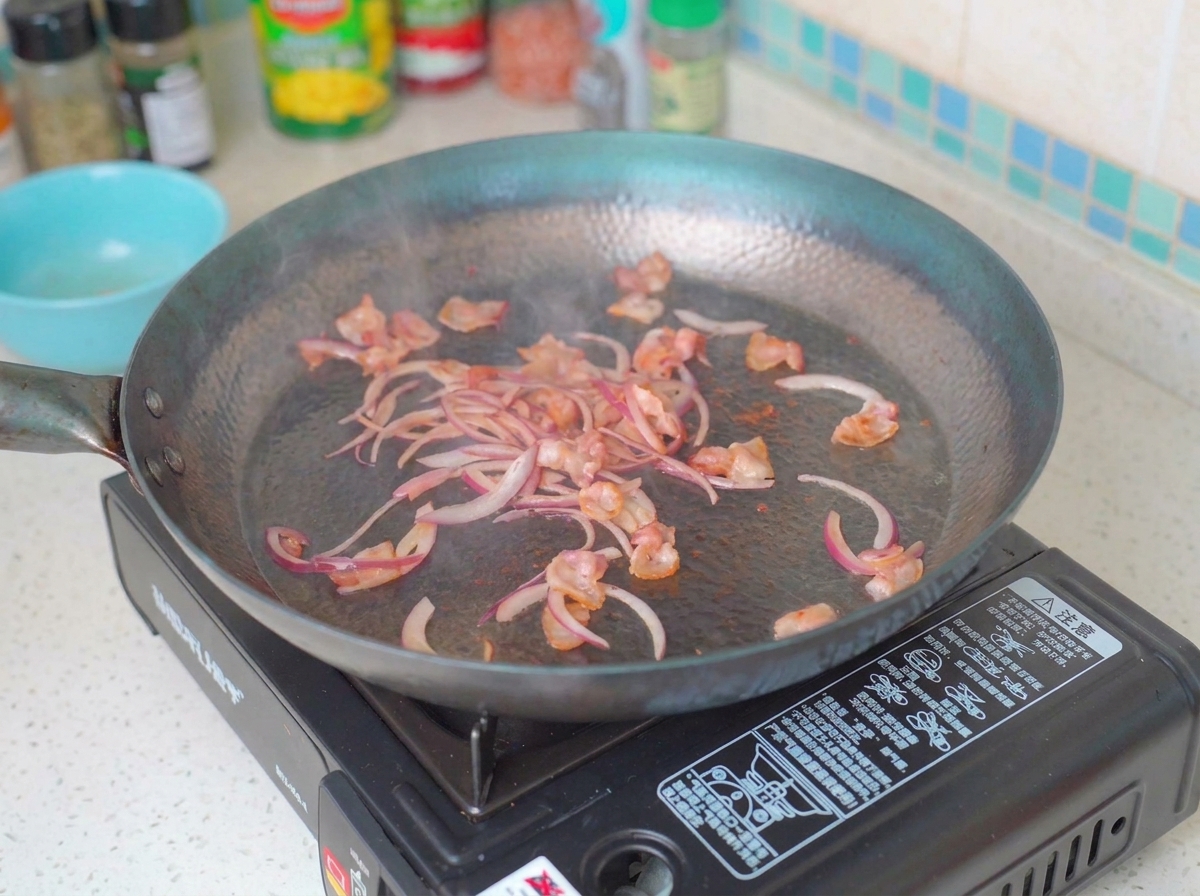 Zwiebelstreifen und Pilze garen in einer Pfanne mit gebratenen Speckstücken.
