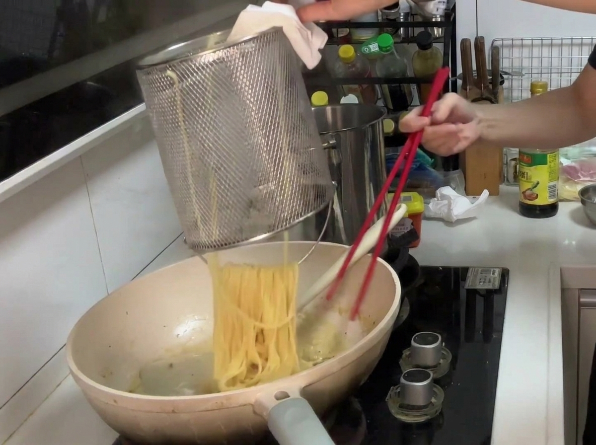 Pasta wird aus einem Sieb direkt in die Pfanne mit der cremigen Pilzsauce gegeben.