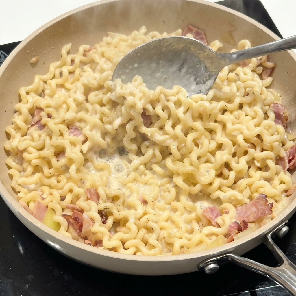 Ein Löffel rührt die Nudeln und den Speck in der cremigen Soße in einer Pfanne um.