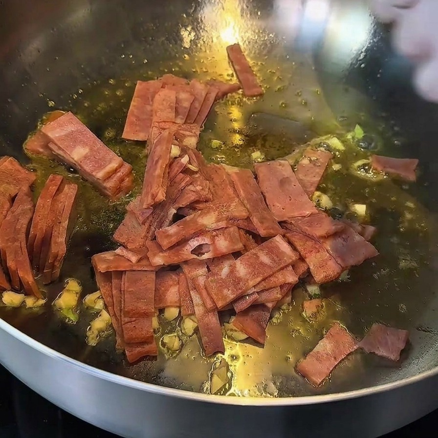 Speck und Knoblauch braten in einer Pfanne, die Ränder des Specks sind bereits goldbraun.