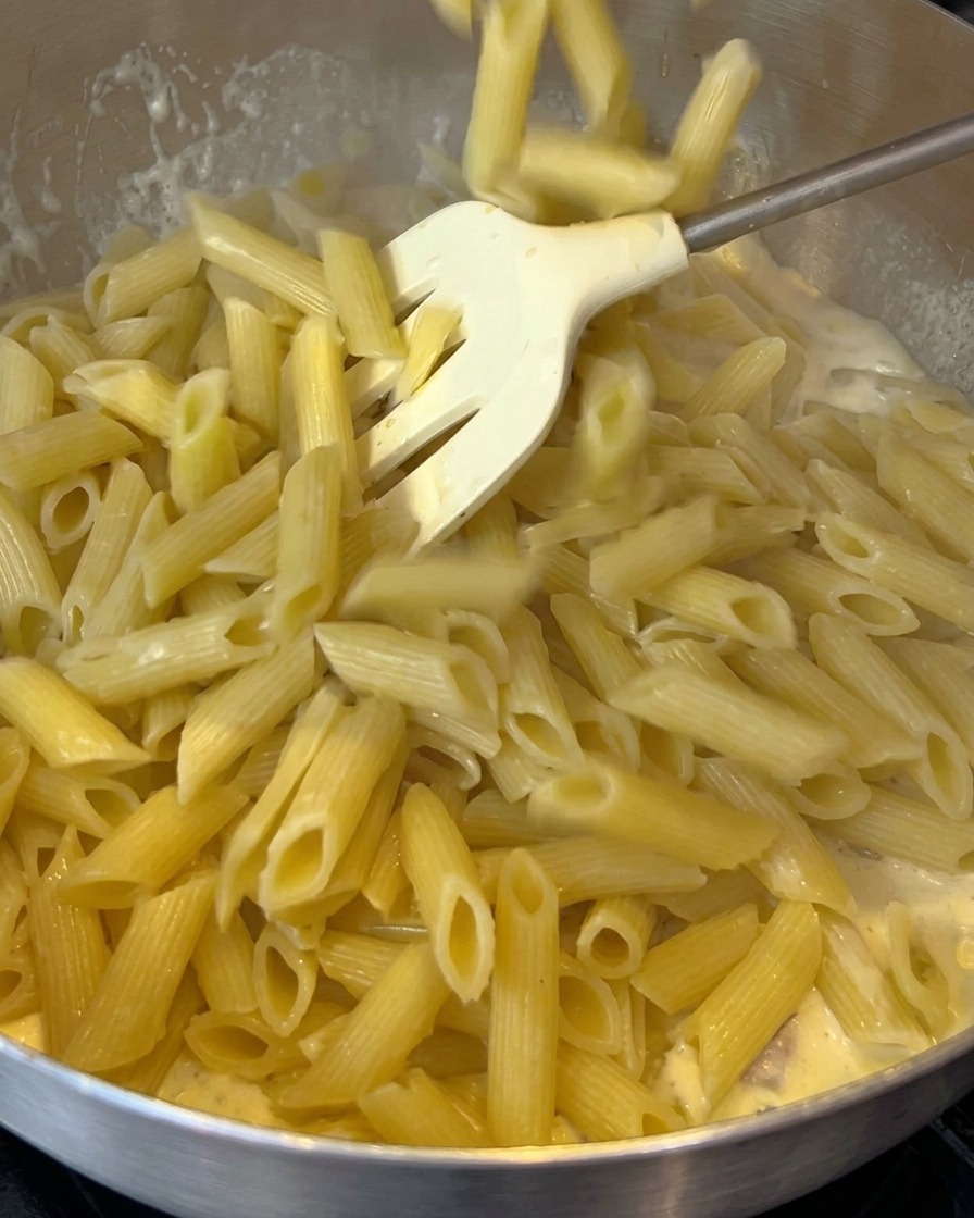 Gekochte Penne werden in eine Pfanne mit cremiger Specksauce gegeben.