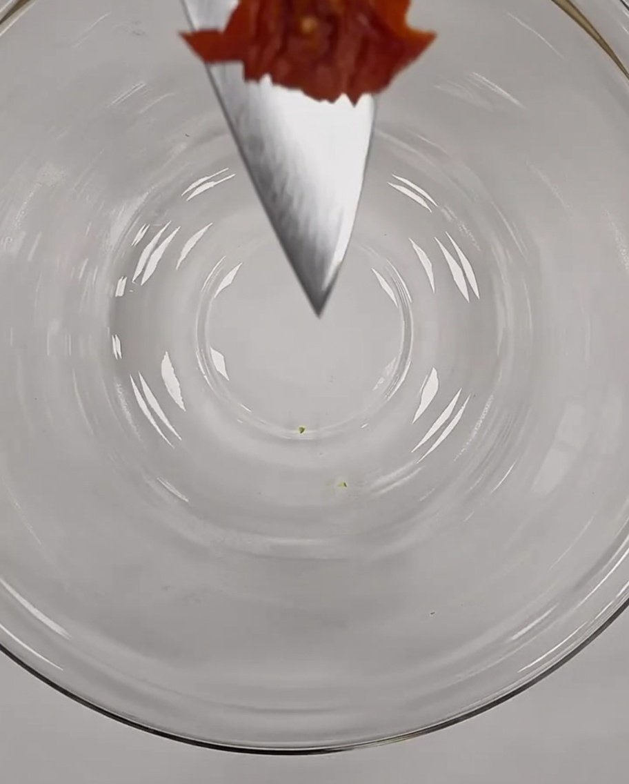 Ein kleines, fein gehacktes Stück getrocknete Tomate auf der Spitze eines Kochmessers über einer Glasschüssel.