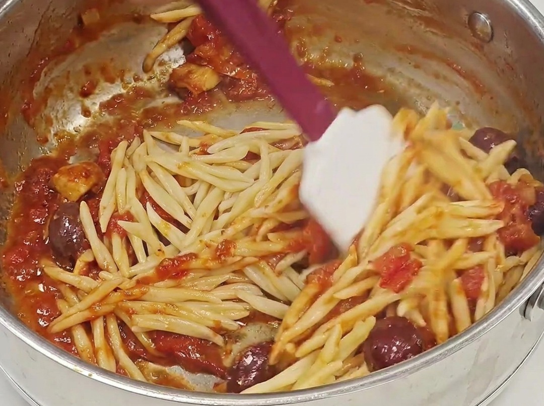 Abgetropfte Trofie-Pasta, die in eine Pfanne mit reichhaltiger, roter Puttanesca-Sauce gegeben wird.