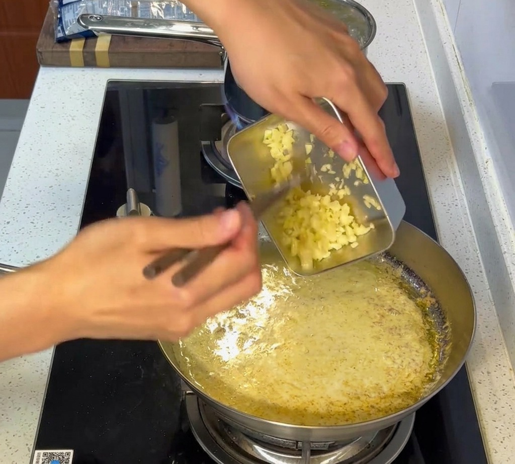 Gehackter Knoblauch wird in eine cremige weiße Sauce in einer Edelstahlpfanne gerührt.