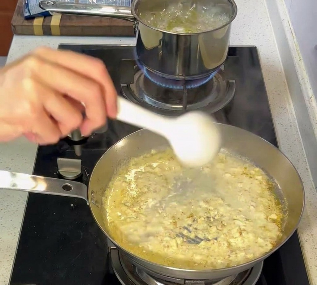Eine Zange überführt al dente gekochte Nudeln aus einem kochenden Topf in die Pfanne mit der Sahnesauce.