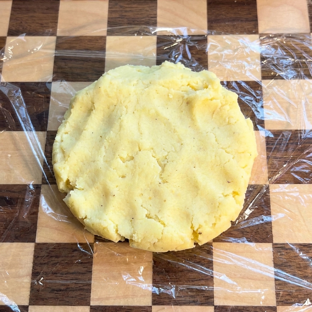 Ein flacher, runder Kartoffelfladen liegt auf einem mit Frischhaltefolie bedeckten Schneidebrett.