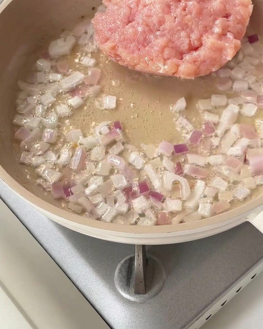 Ein Häufchen rohes gehacktes Hähnchen in einer Pfanne mit angeschwitzten roten Zwiebeln.