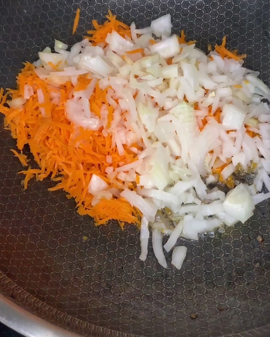 Geriebene Möhren und gewürfelte Zwiebeln dünsten in einer Pfanne bei mittlerer Hitze.