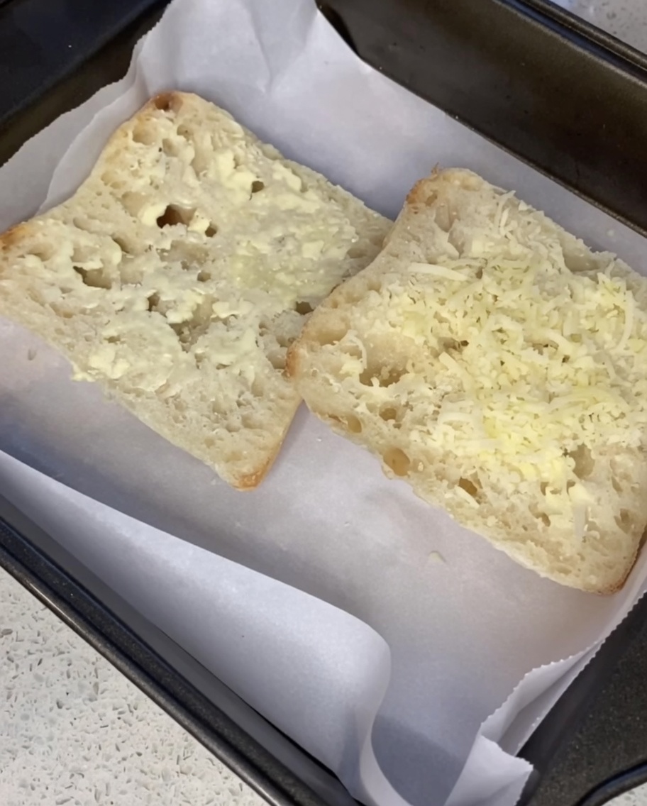Geriebener Parmesan wird über die gebutterten Brotscheiben auf einem Backblech gestreut.