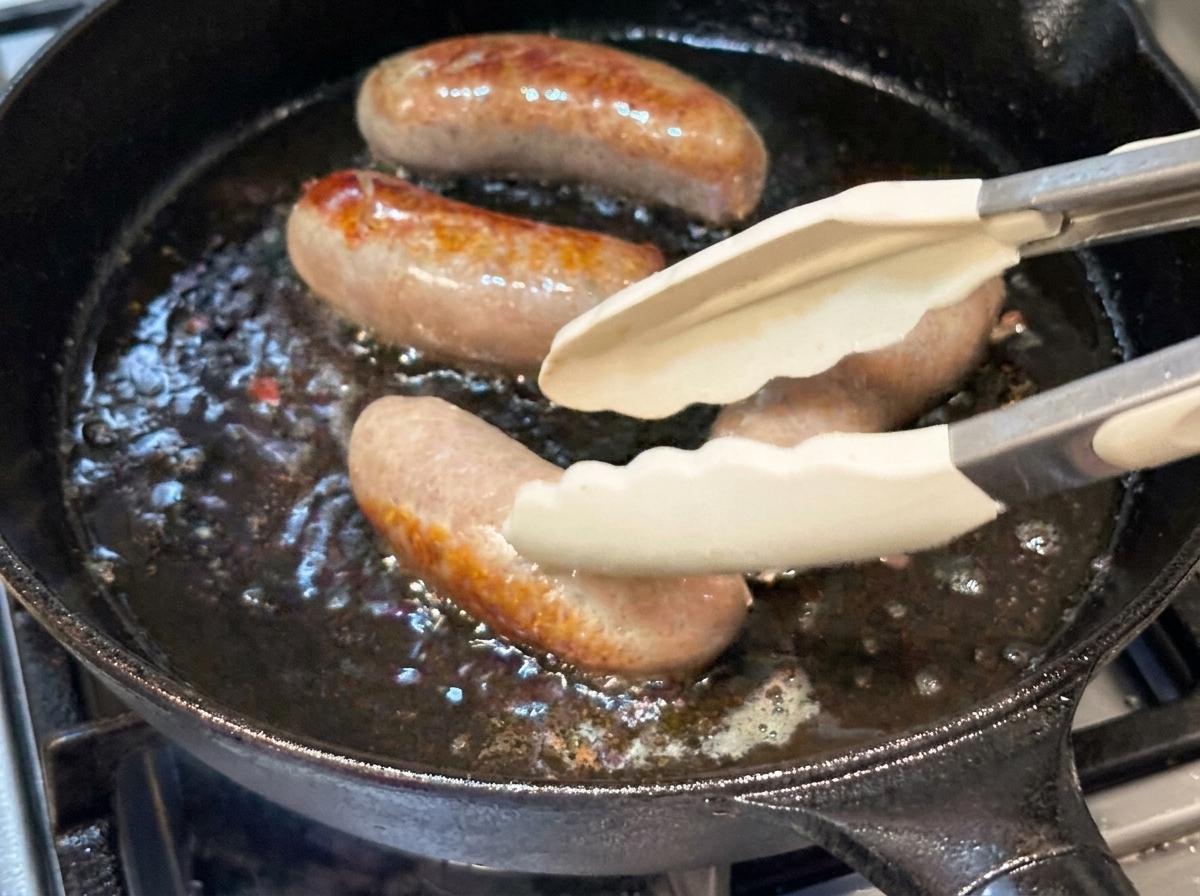 Ganze Würste werden mit einer Silikonzange in einer heißen Gusspfanne gewendet.