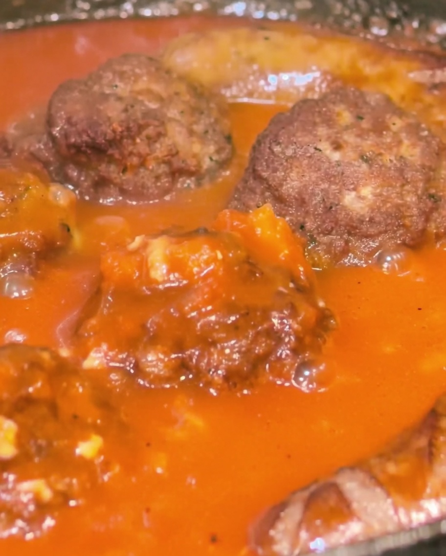 Nahaufnahme einer Pfanne mit Hackbällchen und Würsten, die mit einer dicken, roten Tomatensoße übergossen werden.