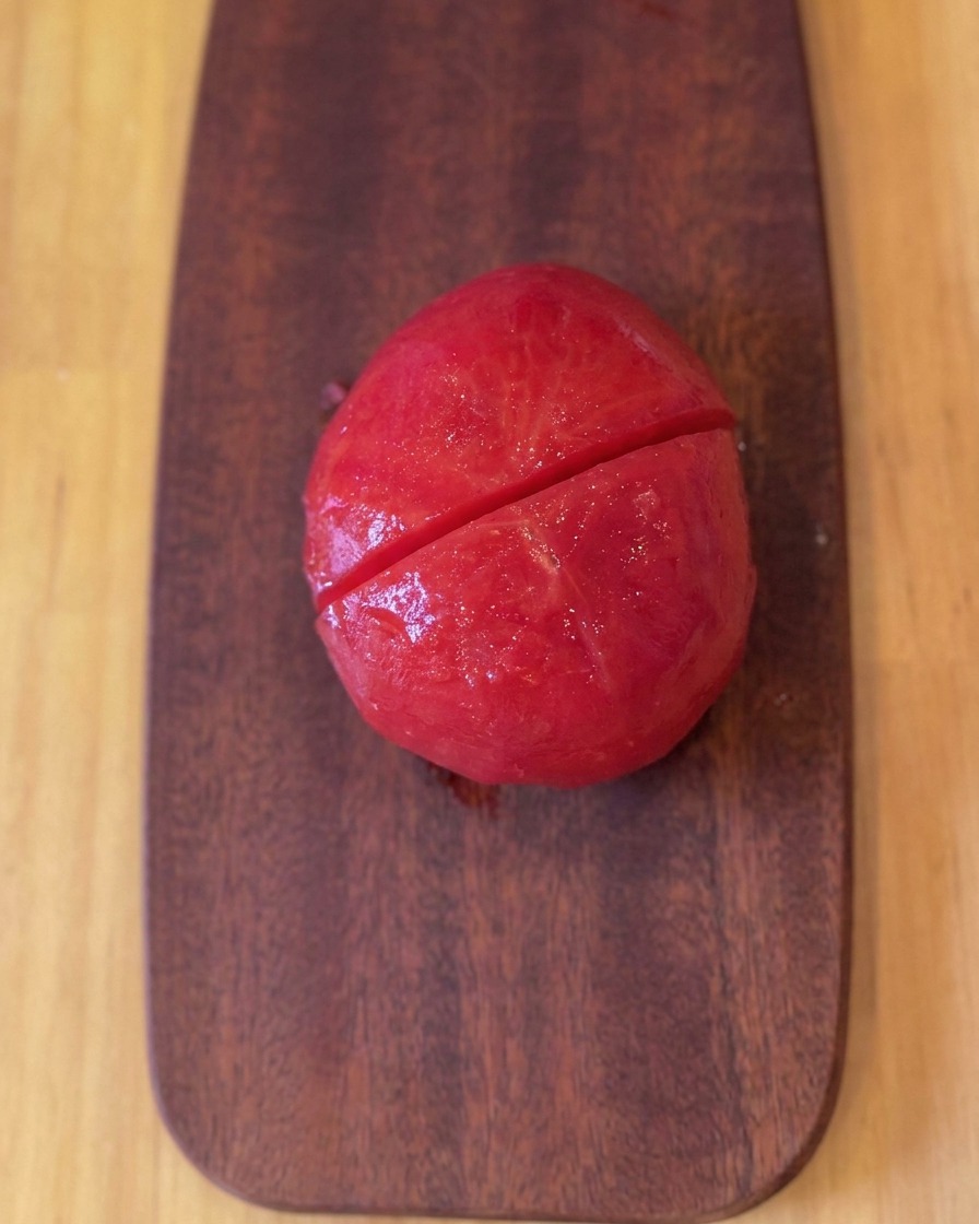 Eine geschälte, geröstete Tomate liegt auf einem Holzbrett und wurde halbiert.