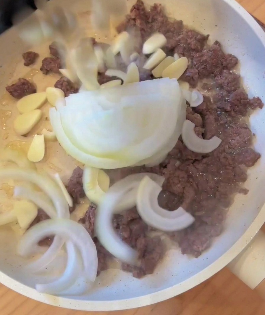 Zwiebeln und Knoblauch werden zum angebratenen Fleisch in die Pfanne gegeben.
