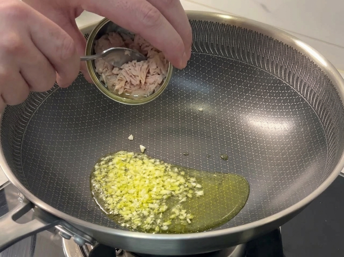 Zarte Thunfischstücke werden mit einem Löffel in eine Pfanne mit brutzelndem Knoblauch gegeben.