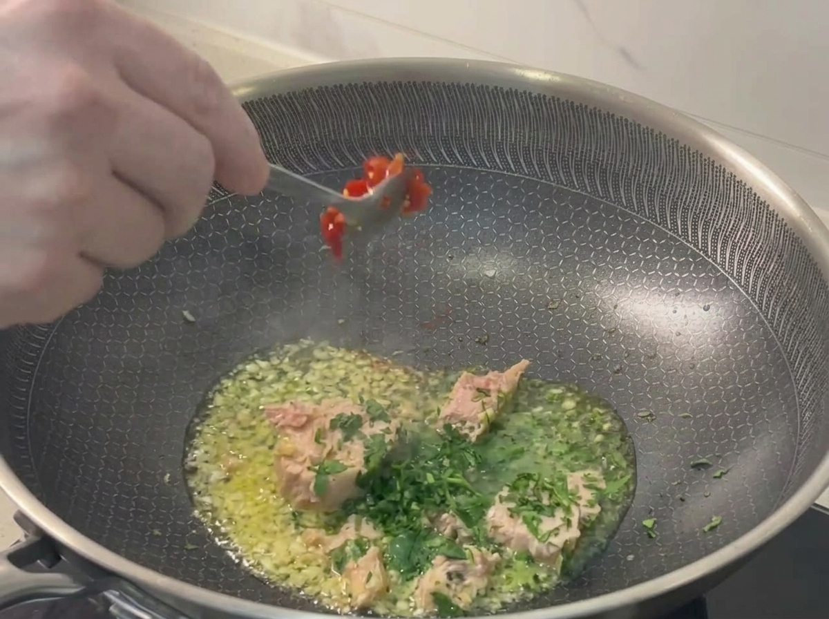 Ein Löffel mit gehackten roten Chilis wird in die Pfanne mit Thunfisch, Knoblauch und Petersilie gegeben.